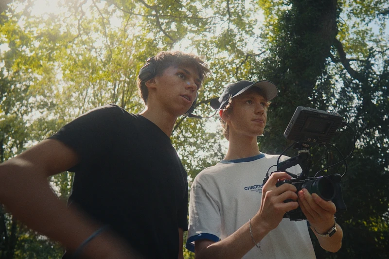 Two young filmmakers with a handheld cinema camera in golden forest light, behind the scenes of a Lua Hell shoot